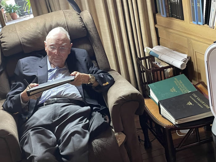Charlie Munger ngả người trên ghế bành đọc tài liệu, cạnh bàn có hai tập Value Line Investment Survey