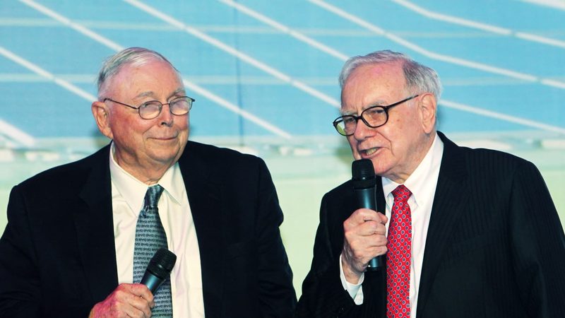 Munger và Buffett cầm micro trên sân khấu, Buffett đang nói